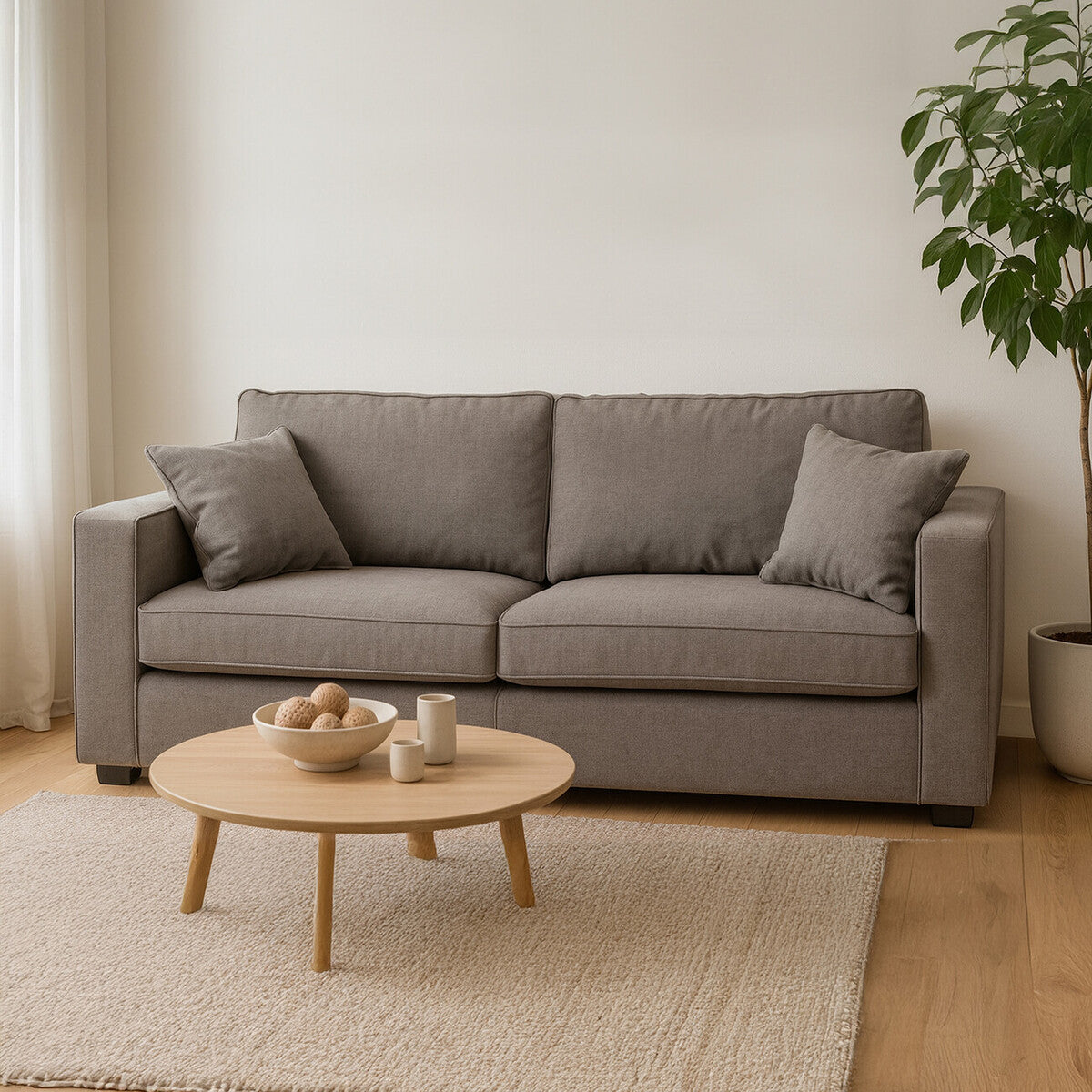 Three-seat grey fabric sofa with two matching cushions, positioned in a bright room with natural light. A round wooden coffee table sits in front of the sofa on a light-colored rug, and a green potted plant is visible in the background