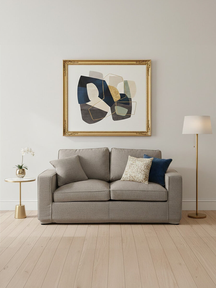A grey two-seat sofa with a modern design, featuring plush cushions and a simple silhouette, positioned in a bright room with light wooden flooring. A decorative throw pillow in navy blue is placed on one side of the sofa. Behind the sofa, there is a large framed abstract artwork with bold colors, and to the left, a stylish floor lamp stands next to a small side table