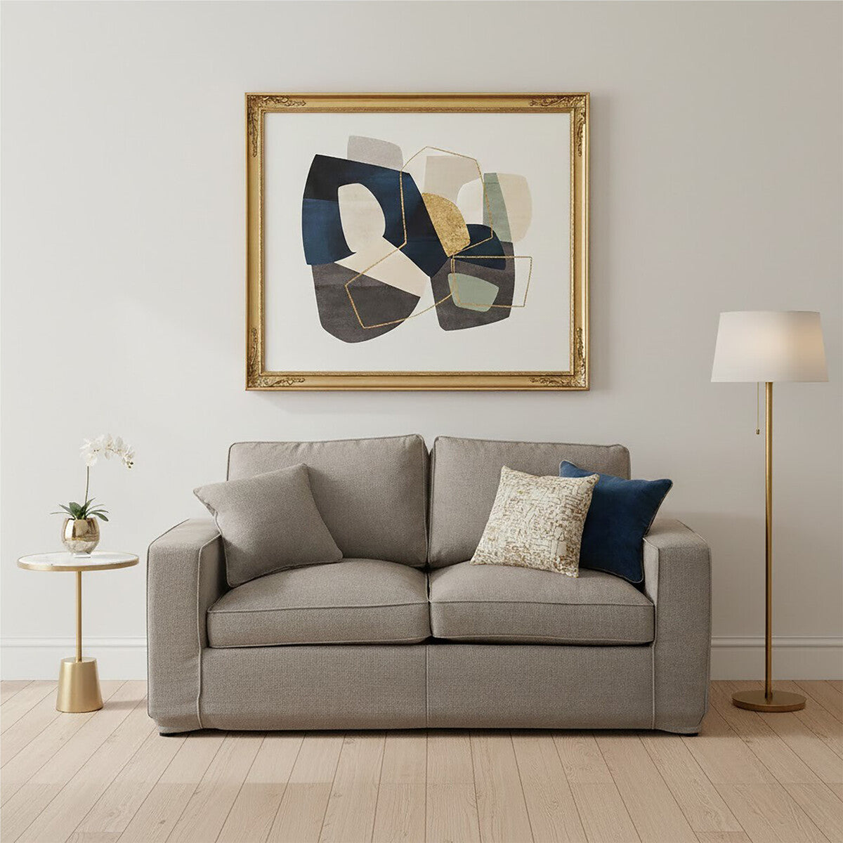 A grey two-seat sofa with plush cushions, positioned against a light-colored wall. A decorative pillow in navy blue and a cream-colored pillow are placed on one side of the sofa. To the left, a small side table holds a vase with flowers, and a floor lamp stands nearby. Above the sofa, an abstract artwork in a gold frame adds a touch of color to the room. The flooring is