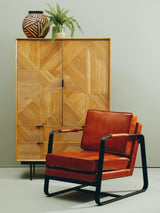 Natural finish cabinet with geometric patterns and two drawers, positioned next to a modern orange armchair with black metal frame, against a neutral wall. A decorative vase and a small plant are placed on top of the cabinet