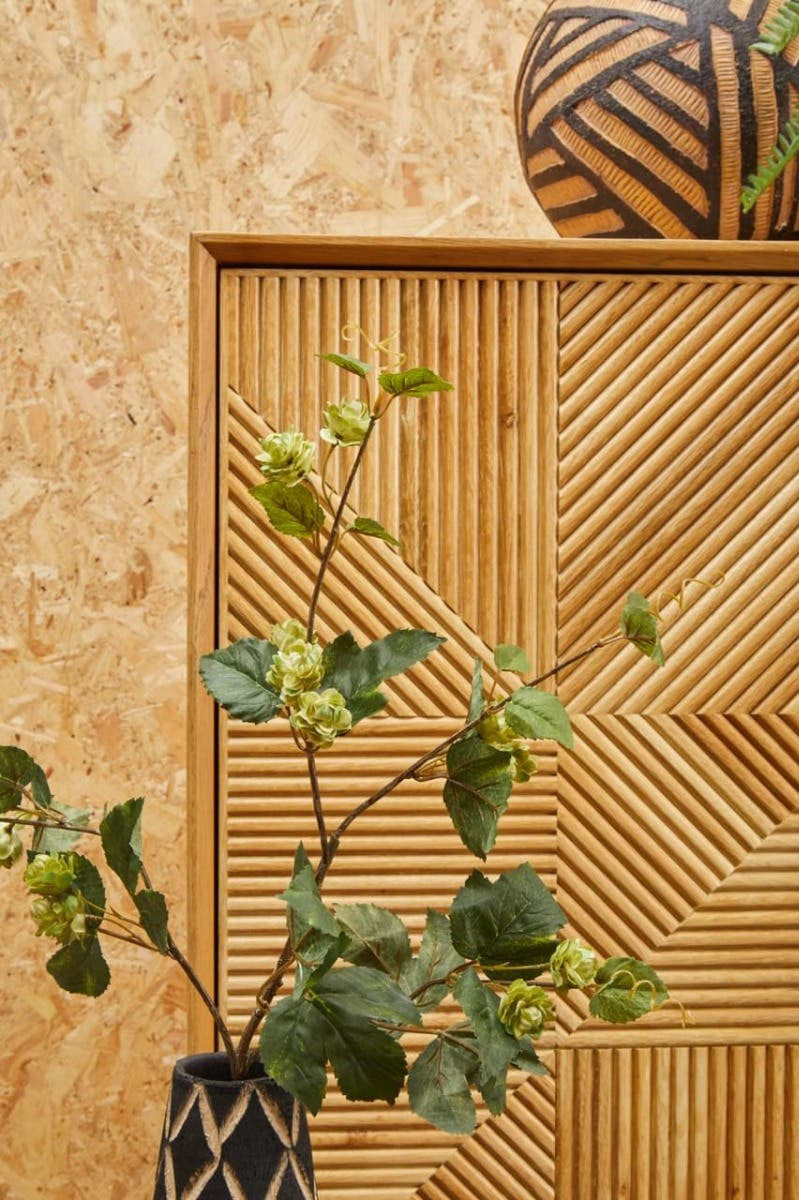 Natural wood cabinet with a textured front, featuring geometric patterns and two drawers. A decorative vase with green foliage is positioned in front of the cabinet, and a patterned pot sits on top of it. The background consists of a light-colored, textured surface