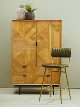 Natural wood cabinet featuring a geometric design, two doors with a central handle, and three drawers at the bottom. A green upholstered chair with a wooden frame is positioned beside the cabinet. The cabinet is topped with two decorative planters. The background is a light, neutral color