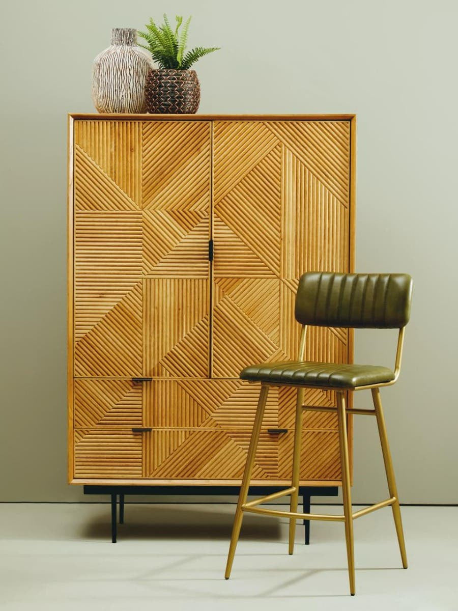 Natural wood cabinet featuring a geometric design, two doors with a central handle, and three drawers at the bottom. A green upholstered chair with a wooden frame is positioned beside the cabinet. The cabinet is topped with two decorative planters. The background is a light, neutral color