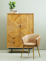 Natural finish cabinet with geometric patterns on the doors, featuring two drawers at the bottom and a potted plant on top. Next to the cabinet, there is a light-colored, upholstered chair with a curved backrest and wooden legs, set against a neutral wall