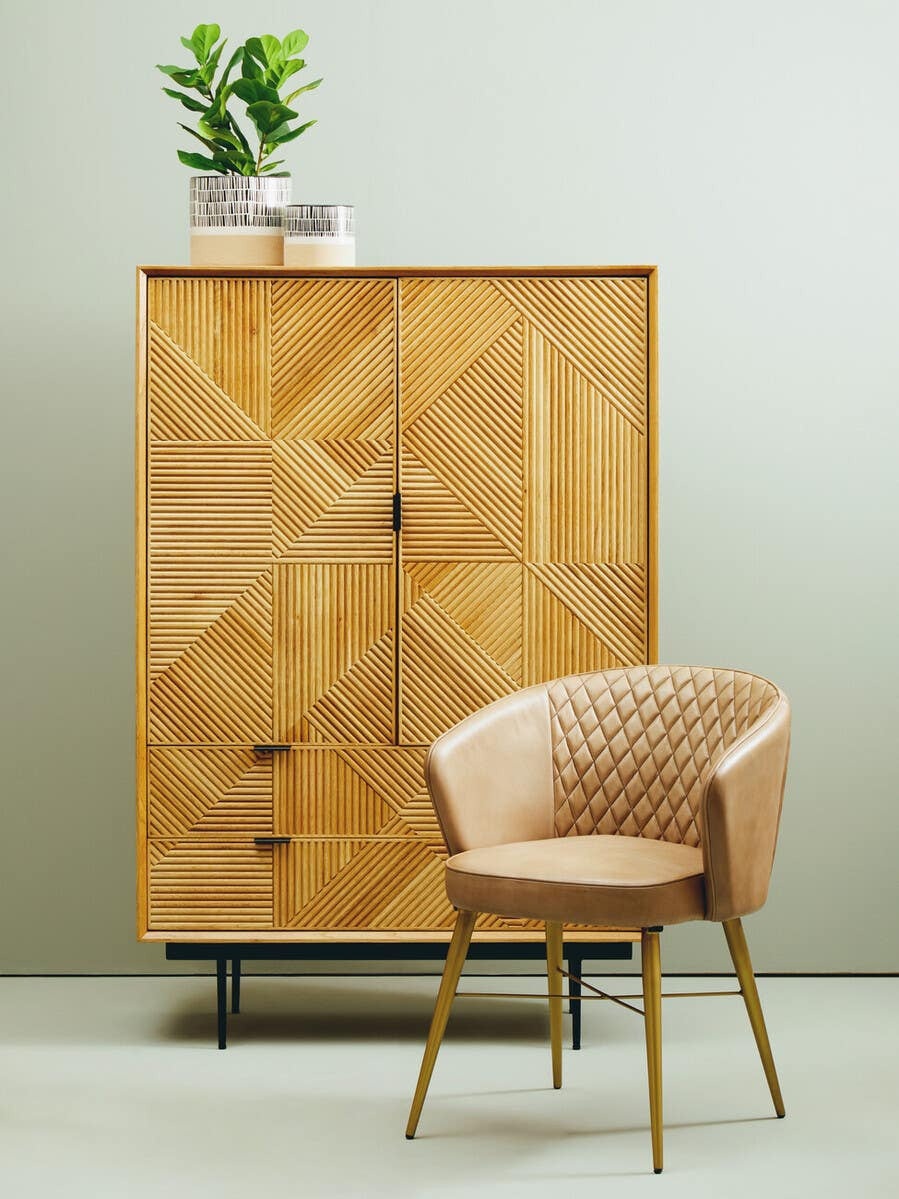 Natural finish cabinet with geometric patterns on the doors, featuring two drawers at the bottom and a potted plant on top. Next to the cabinet, there is a light-colored, upholstered chair with a curved backrest and wooden legs, set against a neutral wall