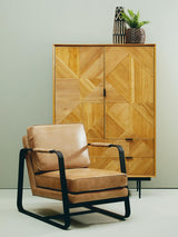 Natural finish cabinet with geometric patterns on the doors, featuring two drawers at the bottom. A stylish, tan leather armchair with a black metal frame is positioned in front of the cabinet. A small potted plant sits atop the cabinet against a light-colored wall