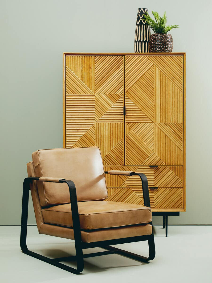 Natural finish cabinet with geometric patterns on the doors, featuring two drawers at the bottom. A stylish, tan leather armchair with a black metal frame is positioned in front of the cabinet. A small potted plant sits atop the cabinet against a light-colored wall