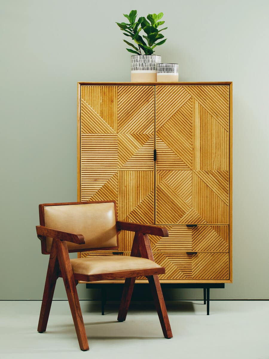 Natural finish cabinet with geometric patterns on the doors, featuring two drawers at the bottom, placed against a light green wall. A small potted plant sits on top of the cabinet. Next to the cabinet, there is a wooden chair with a beige seat and backrest, showcasing a modern design