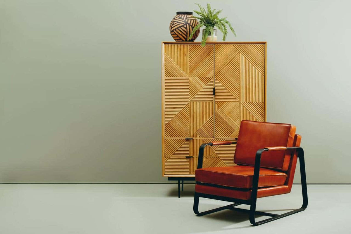 Natural finish cabinet with geometric patterns and two drawers, placed against a light-colored wall. An orange armchair with black metal frame is positioned in front of the cabinet, and a decorative plant sits atop the cabinet alongside a round vase