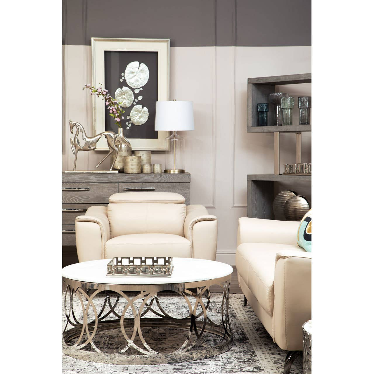 A stylish living room featuring the Padua 3 Seat Stone Leather Sofa with adjustable headrests, complemented by a beige armchair and a round coffee table with decorative items. The backdrop includes a framed artwork and a lamp, creating a cozy and modern atmosphere