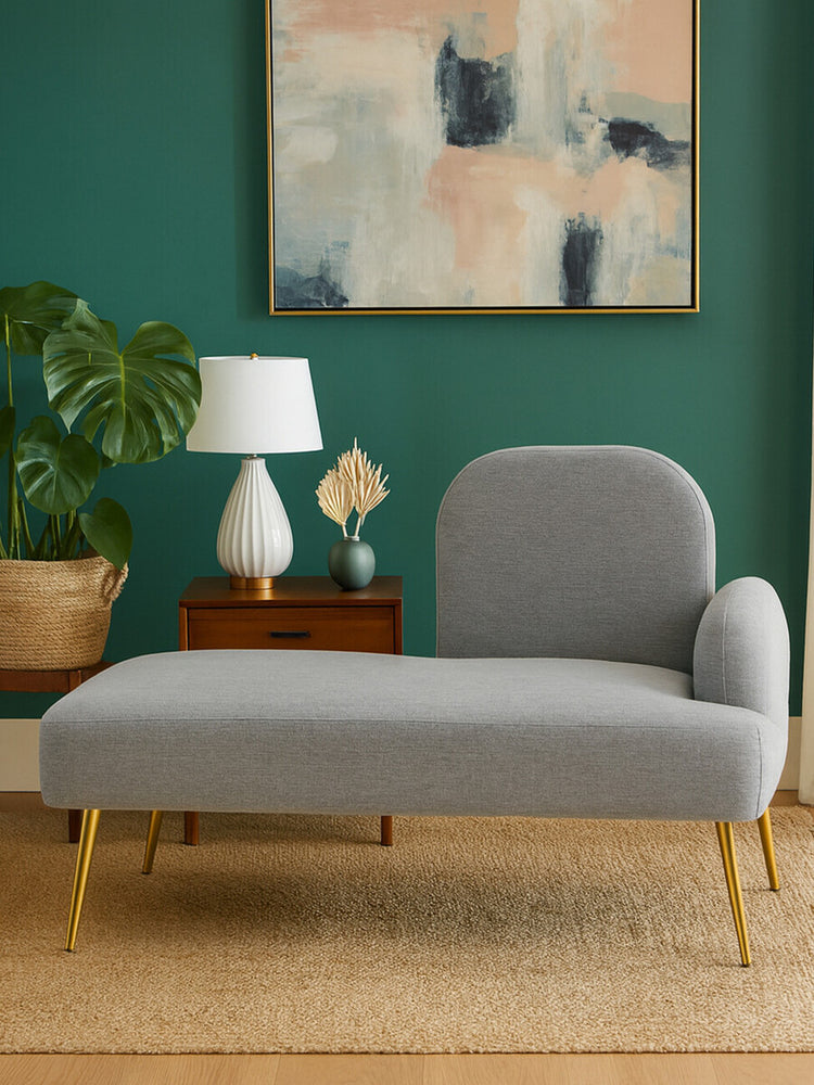 Heather grey chaise longue with a rounded backrest and wooden legs, positioned against a green wall. A white lamp and a small wooden side table with decorative items are visible beside the chaise, along with a potted plant in the background. An abstract painting hangs on the wall above the chaise