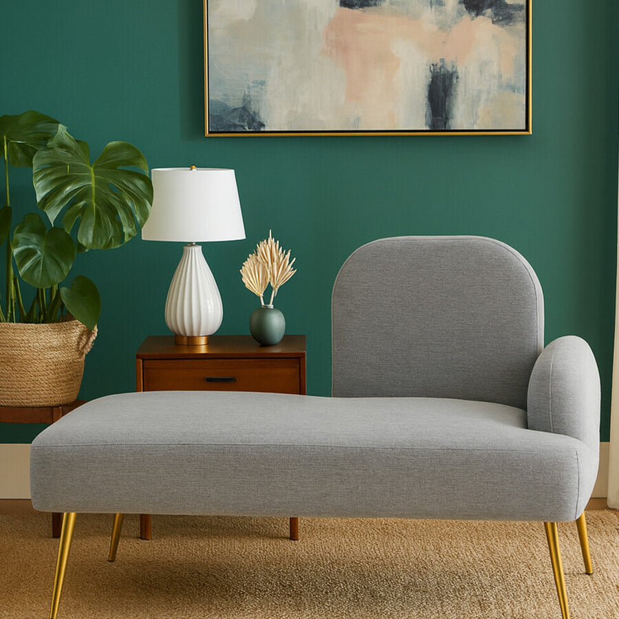 Heather grey chaise longue with a rounded backrest and wooden legs, positioned against a green wall. A white lamp and a small wooden side table with decorative items are visible beside the chaise, along with a potted plant in the background. An abstract painting hangs on the wall above the chaise