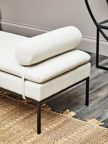 Cream upholstered daybed with black metal legs and bolster cushion on woven rug