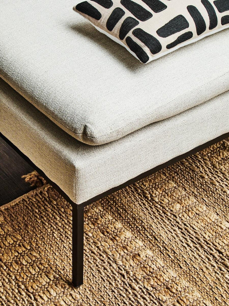 Beige fabric sofa corner with black metal legs, patterned cushion, and woven jute rug