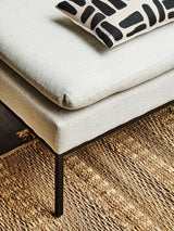 Beige fabric sofa corner with black metal legs, patterned cushion, and woven jute rug