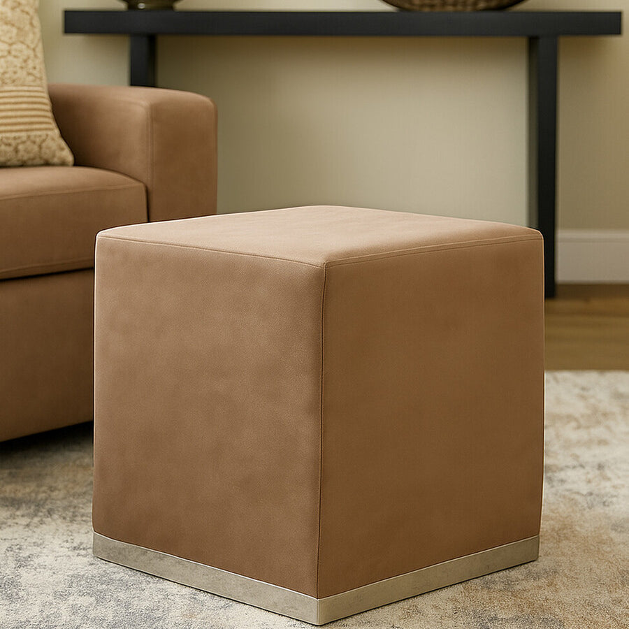 A square stool upholstered in a soft mink-colored fabric with a sleek silver base, positioned on a light-colored rug in a cozy living room setting, next to a brown sofa and a decorative table in the background
