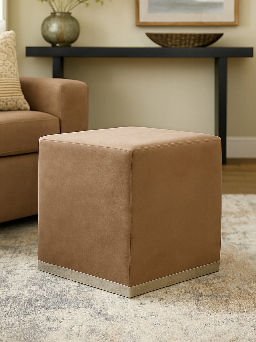 A square stool upholstered in a soft mink-colored fabric with a sleek silver base, positioned on a light-colored rug in a cozy living room setting, next to a brown sofa and a decorative table in the background