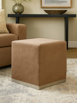 A square stool upholstered in a soft mink-colored fabric with a sleek silver base, positioned on a light-colored rug in a cozy living room setting, next to a brown sofa and a decorative table in the background