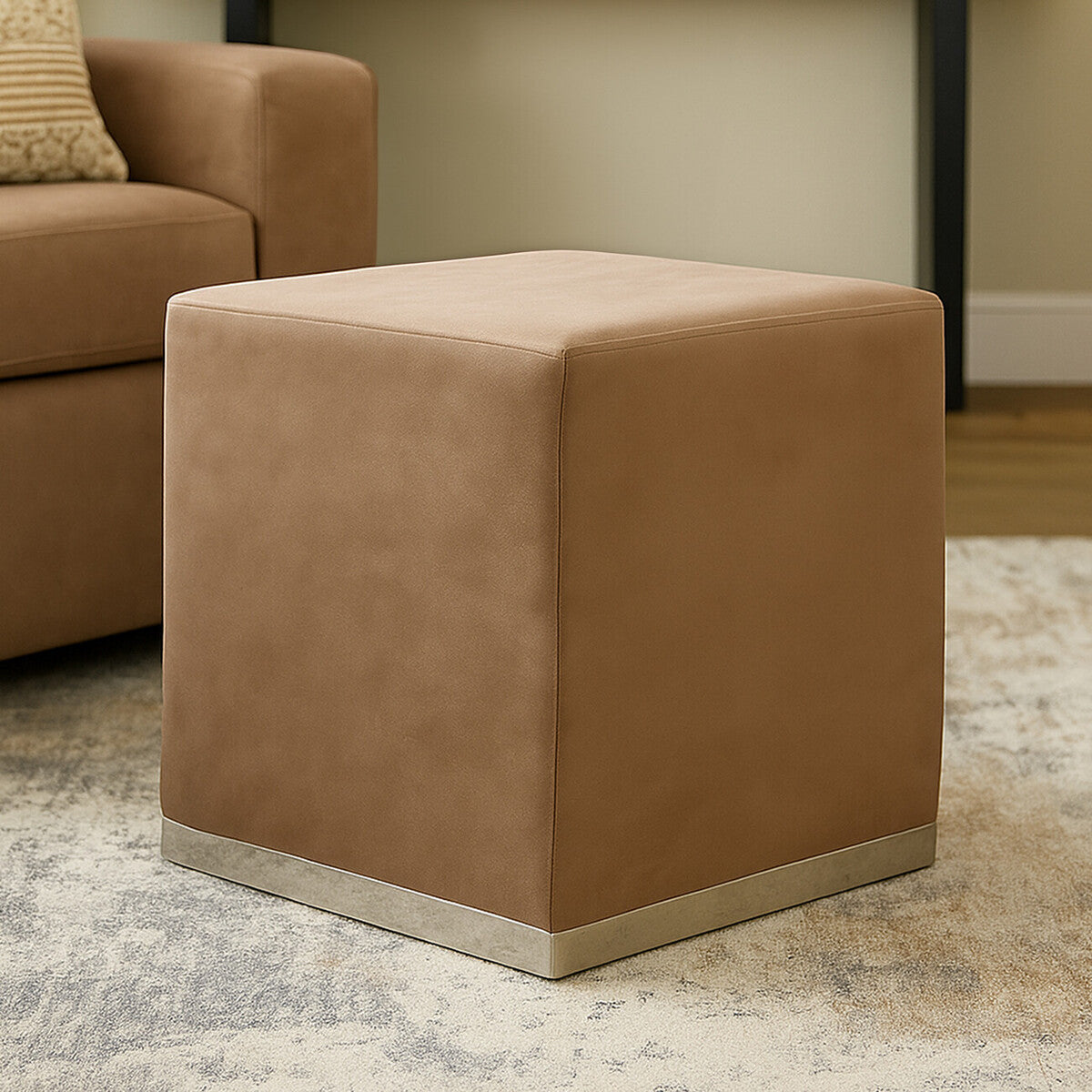 Square stool upholstered in a soft mink-colored fabric with a sleek silver base, positioned on a light-colored rug in a cozy living room setting
