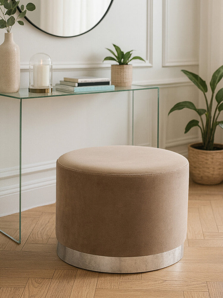 Round stool upholstered in soft mink fabric with a sleek silver base, positioned on a wooden floor near a glass console table. A mirror and potted plants are visible in the background, creating a stylish home decor setting