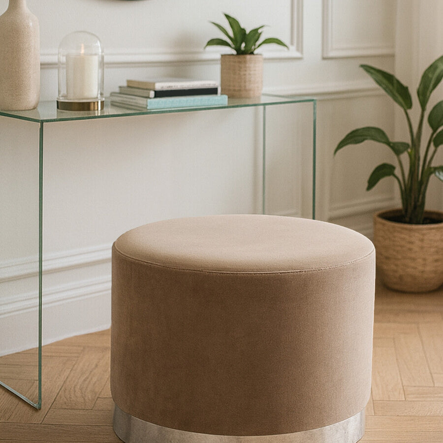 Round stool upholstered in soft mink fabric with a sleek silver base, positioned on a wooden floor near a glass console table. A mirror and potted plants are visible in the background, creating a stylish home decor setting
