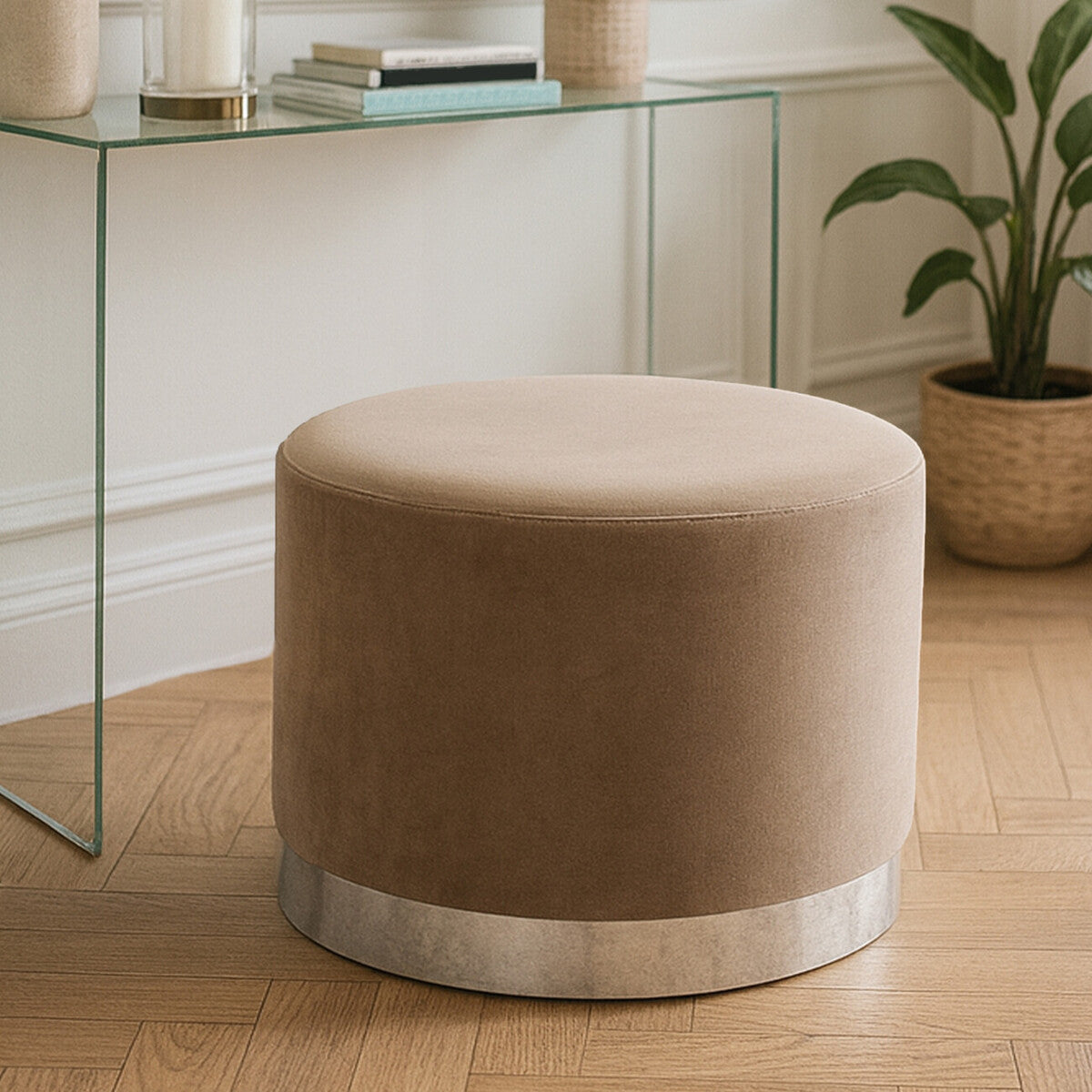 Round stool upholstered in soft mink fabric with a sleek silver base, placed on a wooden floor near a glass table and a potted plant in the background