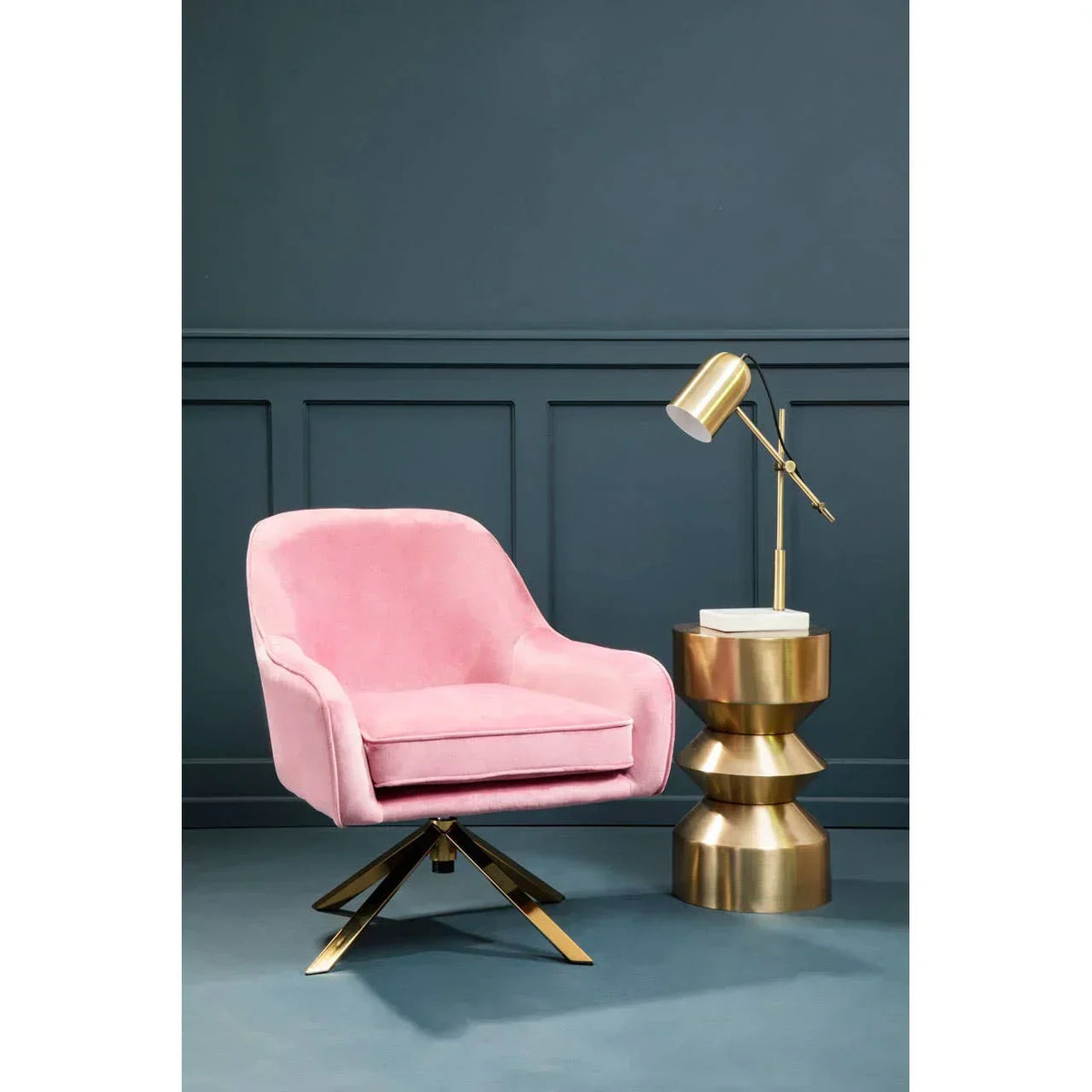Pink velvet accent chair with gold swivel base beside gold side table and desk lamp