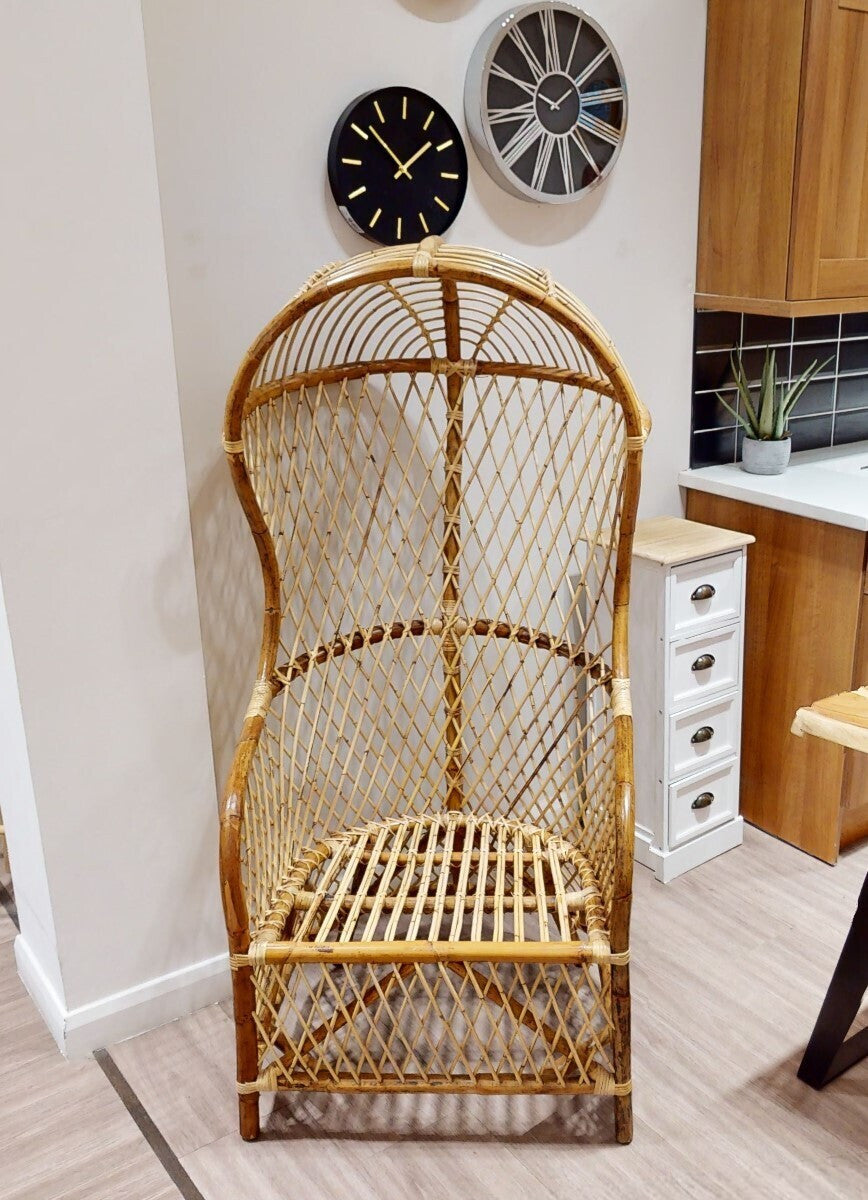 High back natural rattan chair with a woven design, featuring a curved top and open sides, placed in a modern interior setting. Two decorative wall clocks are visible in the background, along with wooden cabinetry and a light-colored floor