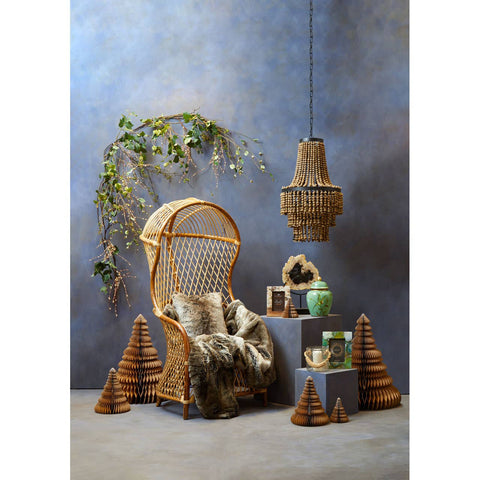 A high back chair made of natural rattan, featuring a curved design and plush cushions, is positioned against a textured blue wall. Nearby, a small round table holds decorative items, including a green vase and a woven basket. A hanging pendant light made of rattan is suspended above, and a leafy plant is draped nearby, adding a touch of greenery to the scene. Three conical decorative pieces