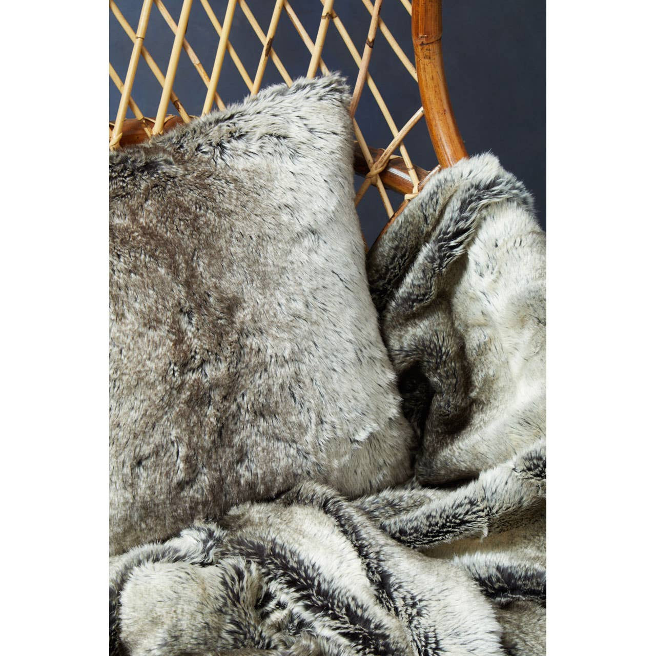 A high back chair made of natural rattan, featuring a woven design and a warm wooden frame. The chair is adorned with a plush, textured throw blanket and a decorative pillow, adding a cozy touch to the seating area. The background is a dark, contrasting color that highlights the chair's natural materials