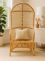 A high back chair made of natural rattan, featuring a woven design and a comfortable seat. It includes a decorative cushion with a subtle pattern, set against a light-colored floor and a bright room with a plant and lamp in the background