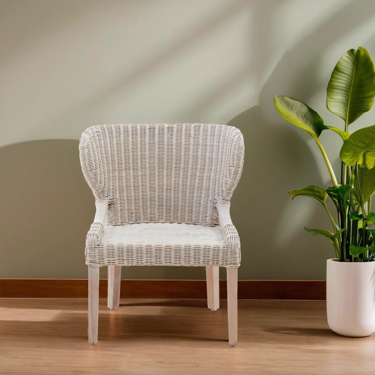 A white wicker chair with a curved backrest and armrests, positioned on a wooden floor against a light green wall, with a potted plant in the background