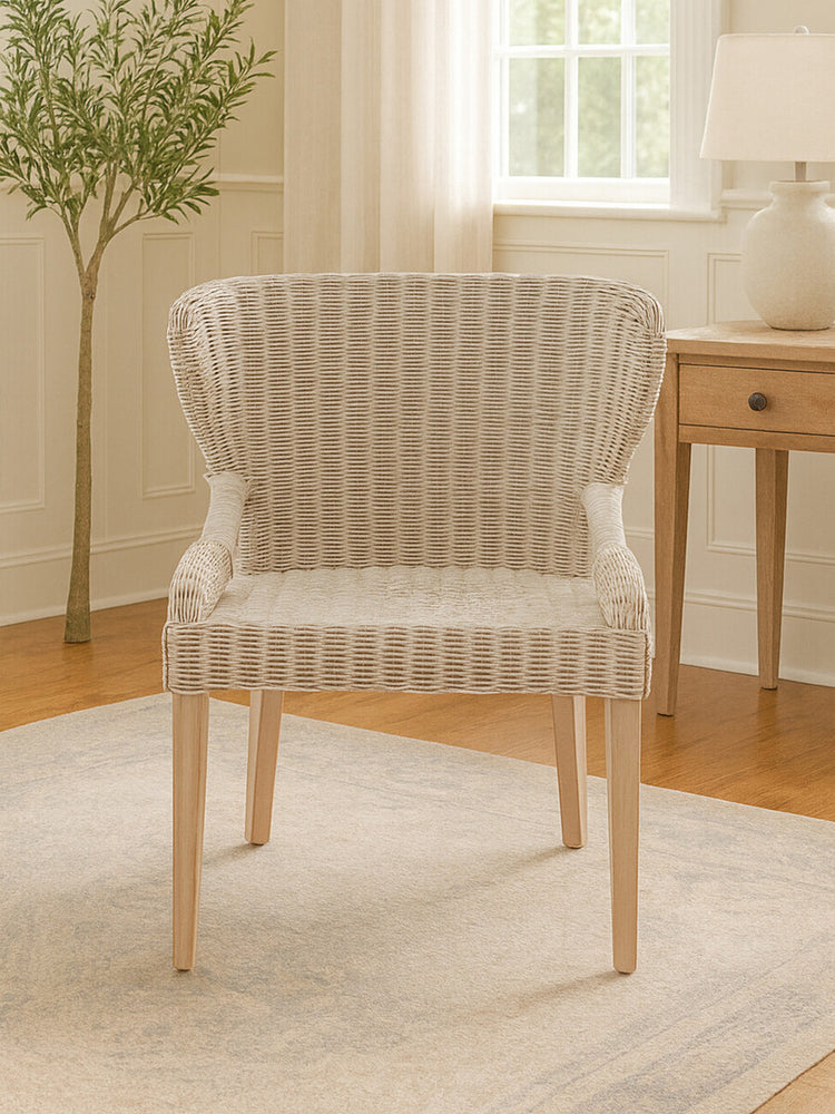 Wicker chair with a curved backrest and light-colored upholstery, featuring wooden legs, placed on a light rug in a bright room with natural light and a small plant in the background
