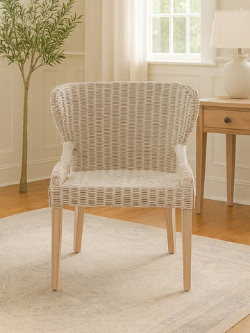Wicker chair with a curved backrest and light-colored upholstery, featuring wooden legs, placed on a light rug in a bright room with natural light and a small plant in the background