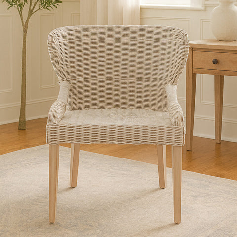 Wicker chair with a curved backrest and armrests, featuring a light beige woven pattern, supported by natural wood legs, placed on a light-colored rug in a cozy room setting with a plant and a wooden side table in the background