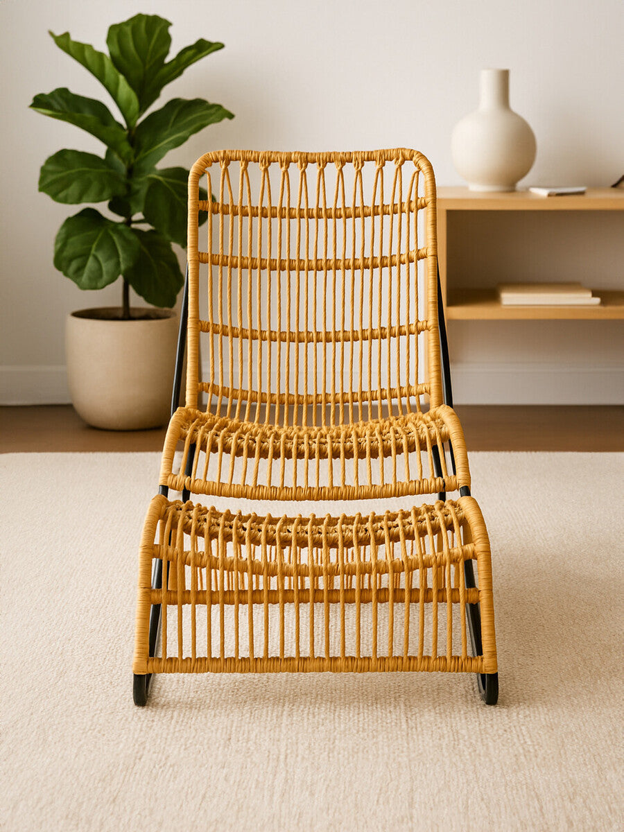 A rattan chair with a natural finish and a matching footstool, featuring a slatted design and a comfortable, relaxed seating position. The chair is positioned on a light-colored carpet, with a green potted plant and a wooden shelf in the background