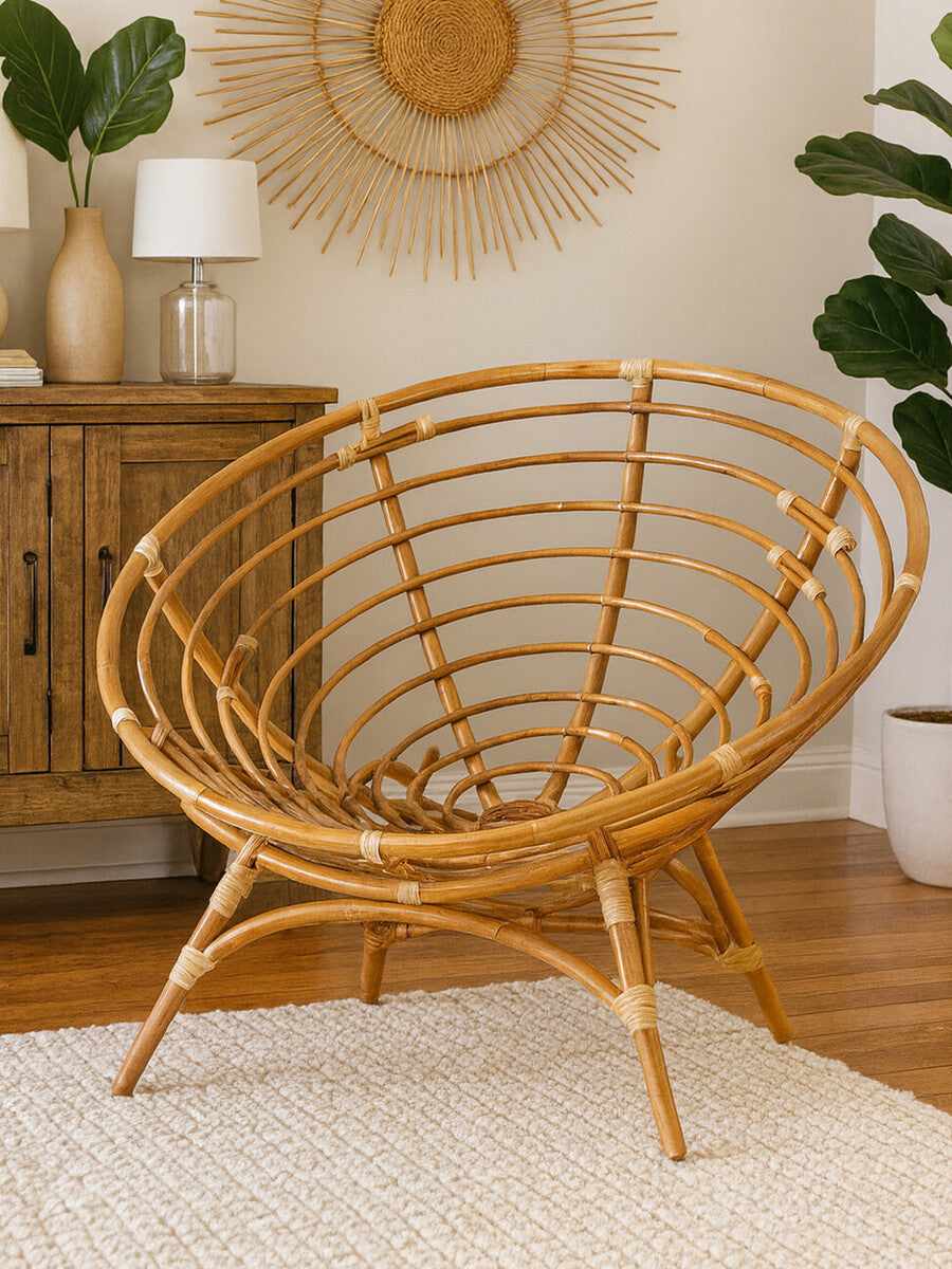 Natural rattan Papasan chair with a circular, open-weave design and a sturdy base, placed on a light-colored rug in a cozy room. A wooden cabinet and a decorative wall hanging are visible in the background, along with a lamp and a potted plant