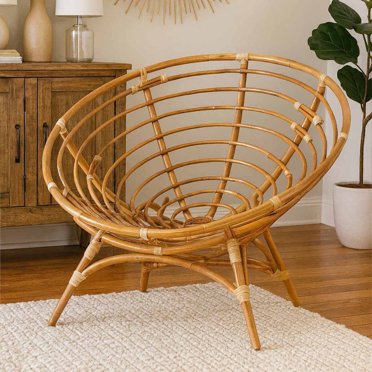 Natural rattan chair with a circular, open-weave design and sturdy legs, positioned on a light-colored rug in a cozy room setting. A wooden cabinet and a potted plant are visible in the background, adding to the warm ambiance