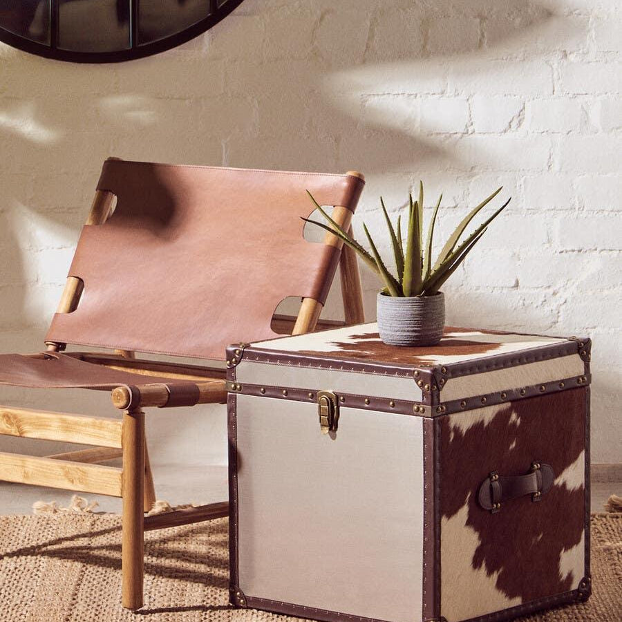 Cowhide storage trunk with brass hardware beside leather accent chair on woven rug