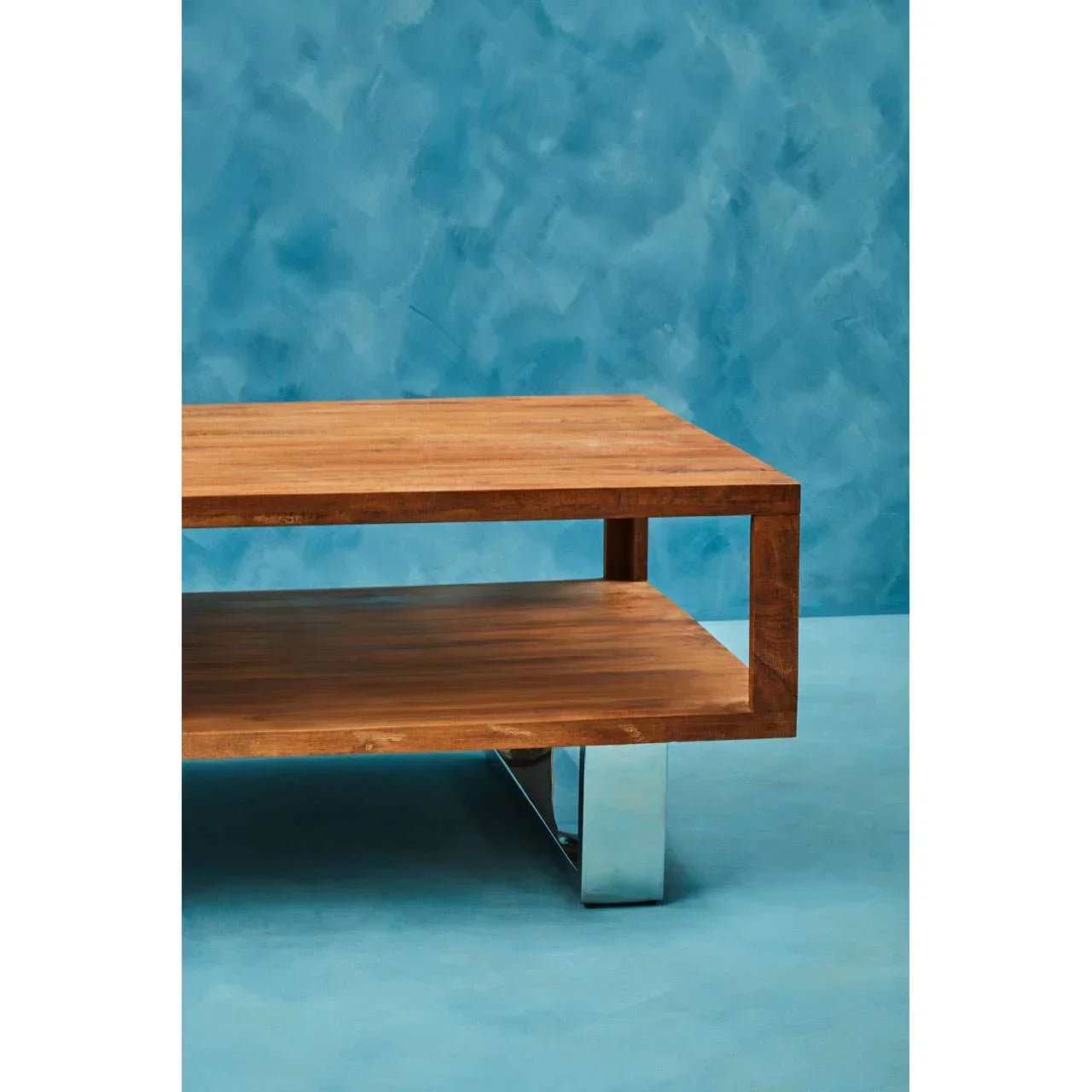 Rectangular wooden coffee table with metal leg and open shelf against blue background