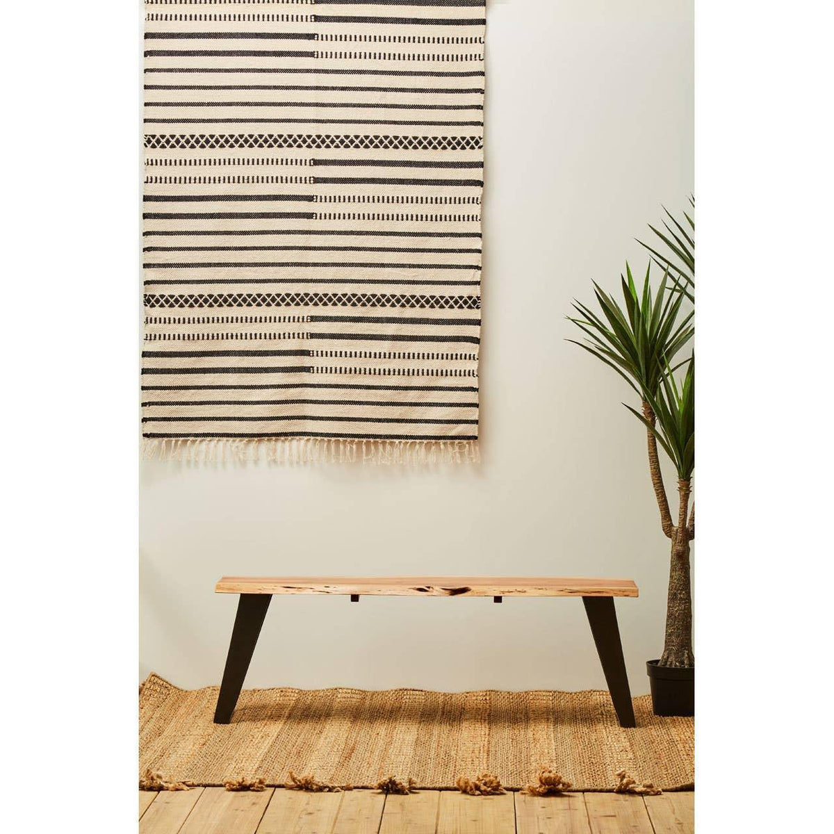 Wooden bench with black legs on woven rug next to potted plant and striped wall hanging