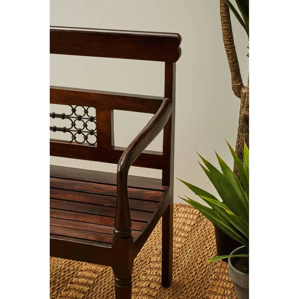 Dark wood bench with decorative backrest and armrest beside indoor plant on woven rug
