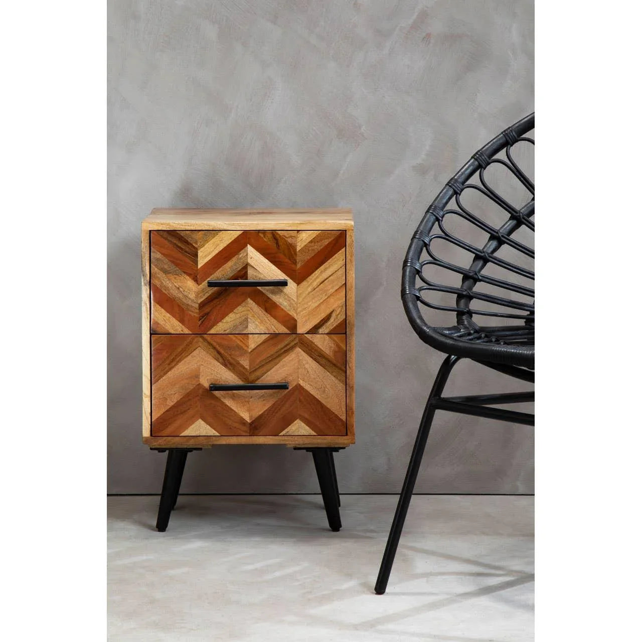 Chevron wood bedside table with two drawers and black handles, next to rattan chair