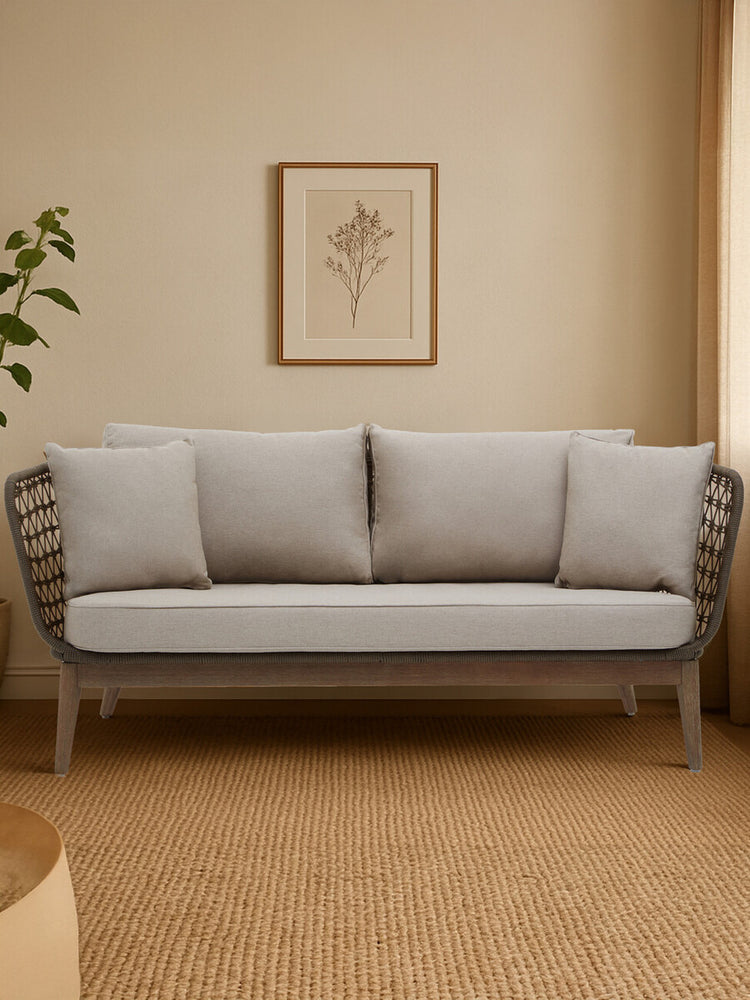 Three-seater sofa featuring a grey woven rope design with plush cushions in a cozy living space. The sofa is positioned against a light-colored wall, complemented by a decorative wall art piece and a potted plant nearby. The flooring is a warm, neutral tone, enhancing the inviting atmosphere