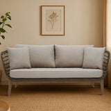 Three-seater sofa featuring a grey woven rope design with a sturdy frame, complemented by plush light grey cushions. The sofa is positioned against a neutral-colored wall, with a framed botanical print above it and a potted plant to the side, set on a warm-toned carpet