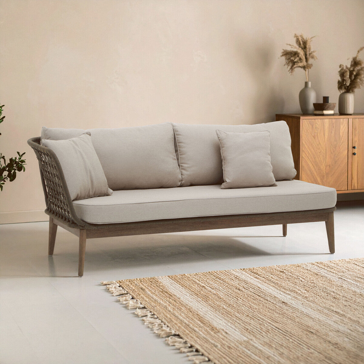 The Opus Woven Rope Chaise Longue features a sleek design with a light beige fabric upholstery and three plush cushions. It has a sturdy wooden frame with a natural finish, complemented by woven rope detailing on the sides. The chaise is positioned on a light-colored floor, with a textured area rug underneath, and a wooden cabinet with decorative items in the background