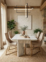 A stylish dining area featuring the Vieste Mink Velvet Dining Chair, which has a soft, plush velvet upholstery in a mink color. The chair is positioned around a modern white marble dining table with a sleek design. Natural light streams in through large windows, illuminating the space, which includes decorative plants and a contemporary light fixture hanging above the table. The floor is covered with a textured rug, adding warmth to