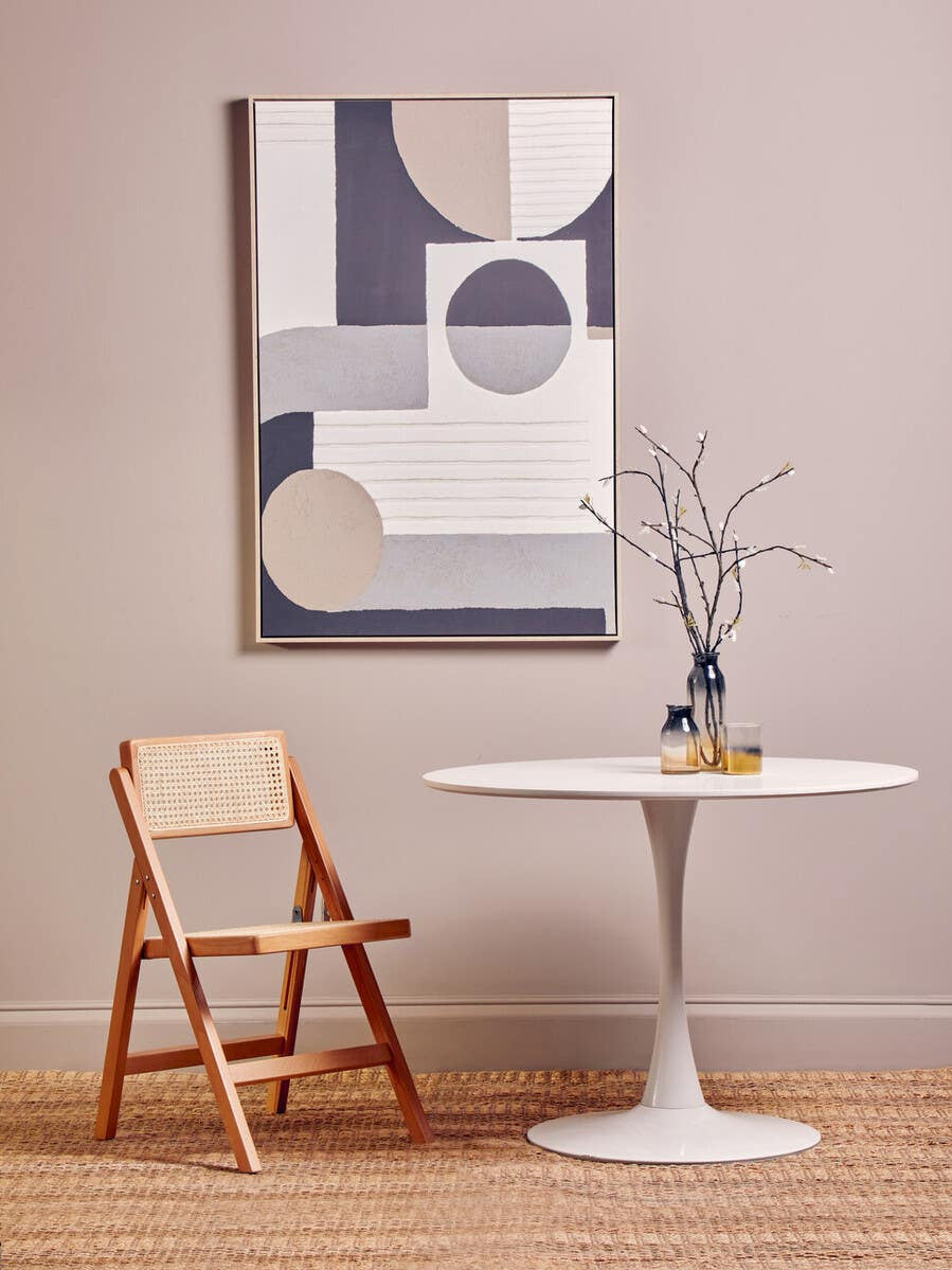 A light-colored beech wood folding chair is positioned next to a round white table. The chair is set against a soft beige wall, and a modern abstract artwork featuring geometric shapes in muted colors hangs above the table. A small vase with a single flower sits on the table, adding a touch of elegance to the scene. The floor is made of light wood, enhancing the warm ambiance of the setting