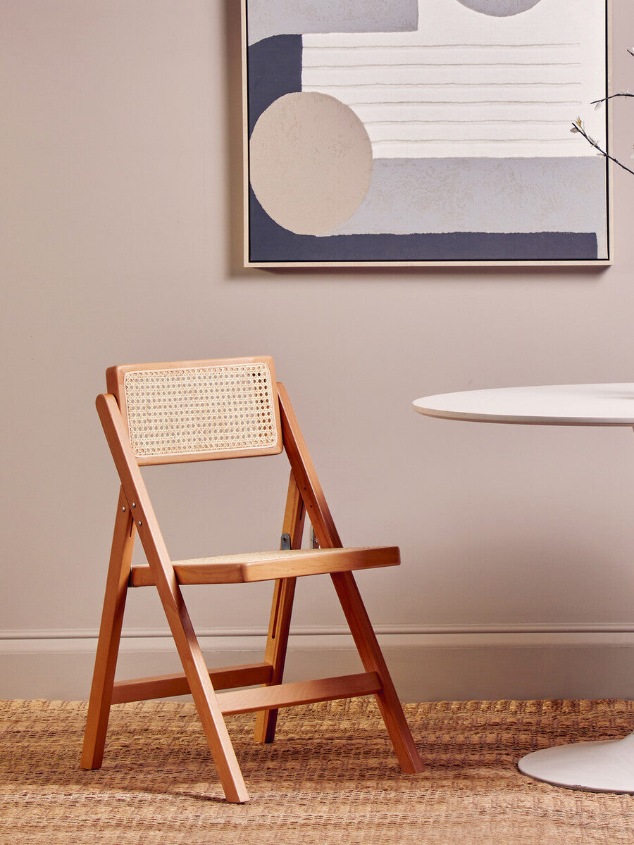 Folding chair made of beech wood with a natural finish, positioned next to a round table on a light-colored floor, against a wall featuring abstract artwork in muted tones