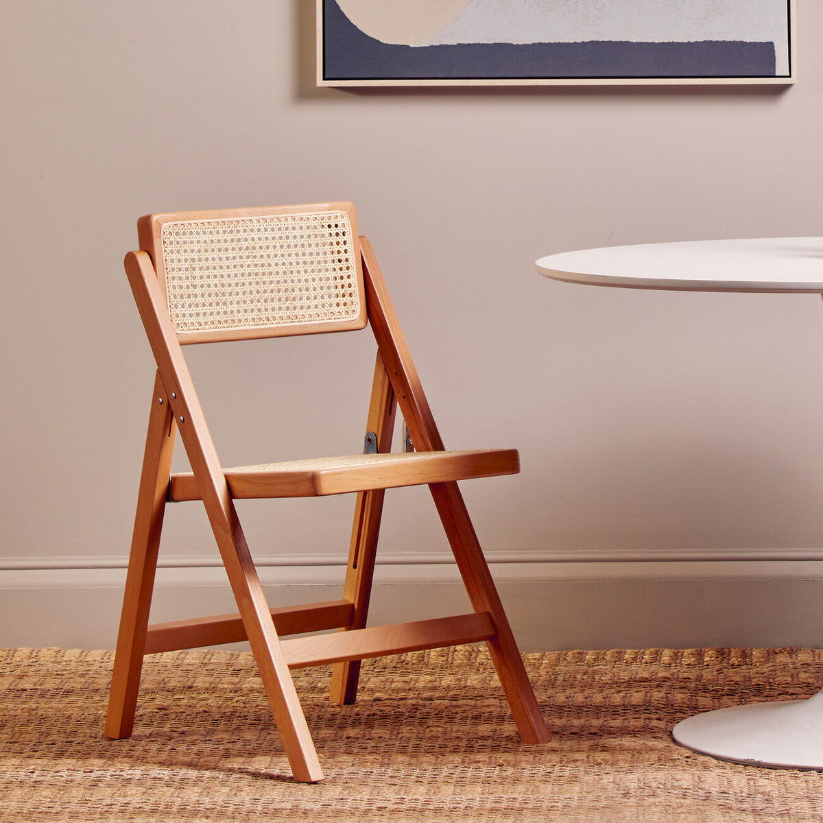 Folding chair made from beech wood with a natural finish, featuring a woven cane backrest and a simple, modern design. The chair is positioned next to a round table on a textured rug, set against a neutral wall with a piece of abstract art above